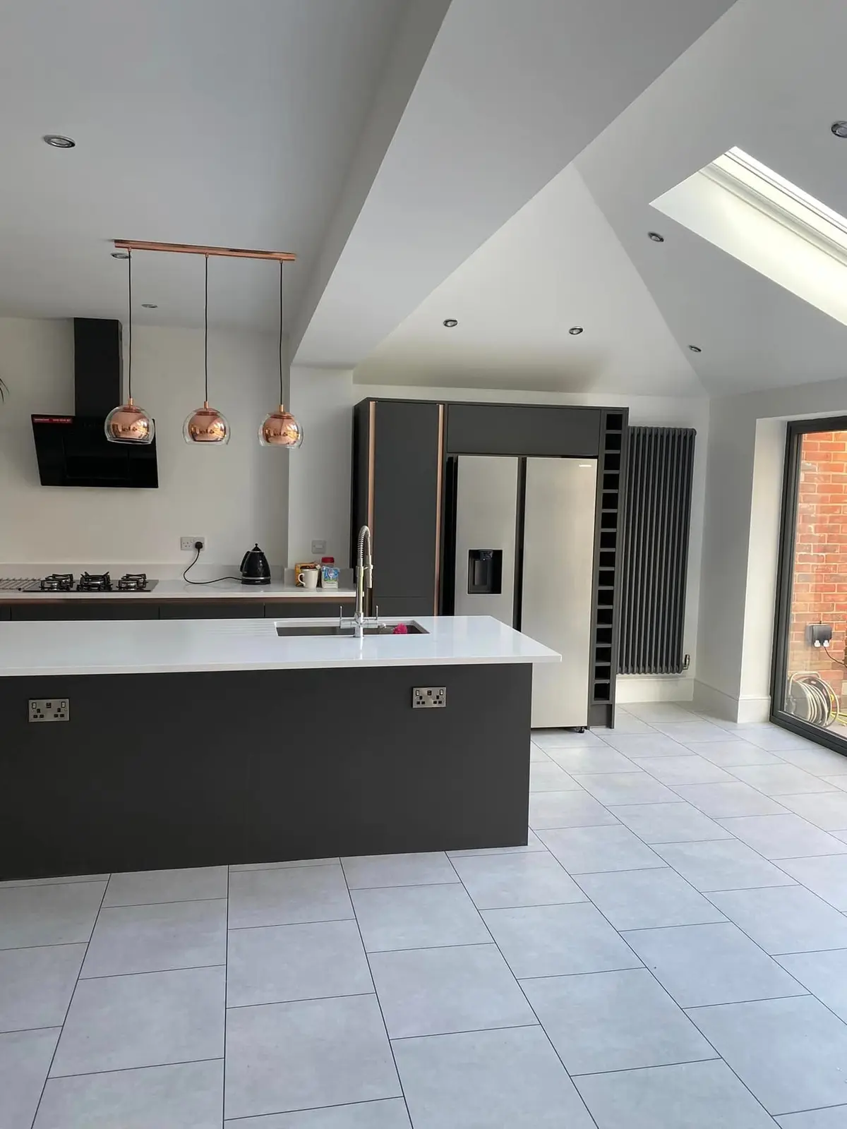 Dark grey handleless kitchen with a long white-topped island and copper pendant lights under a vaulted ceiling with roof windows