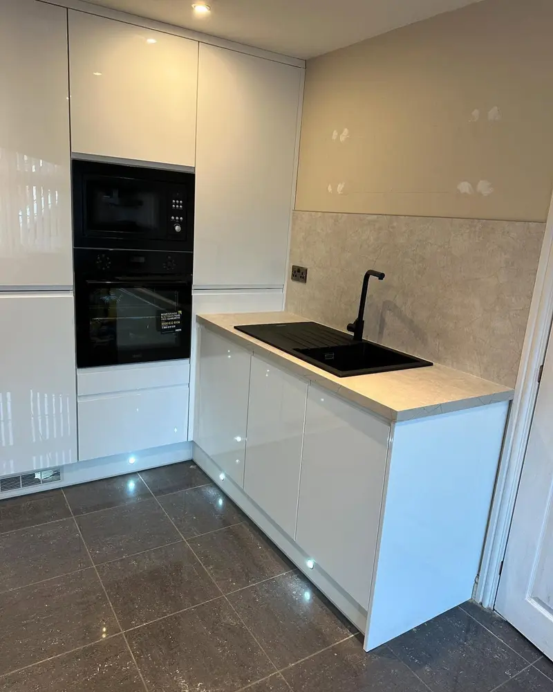 White high-gloss handleless kitchen with tall housing, black built-in ovens, stone-effect worktop and plinth lights